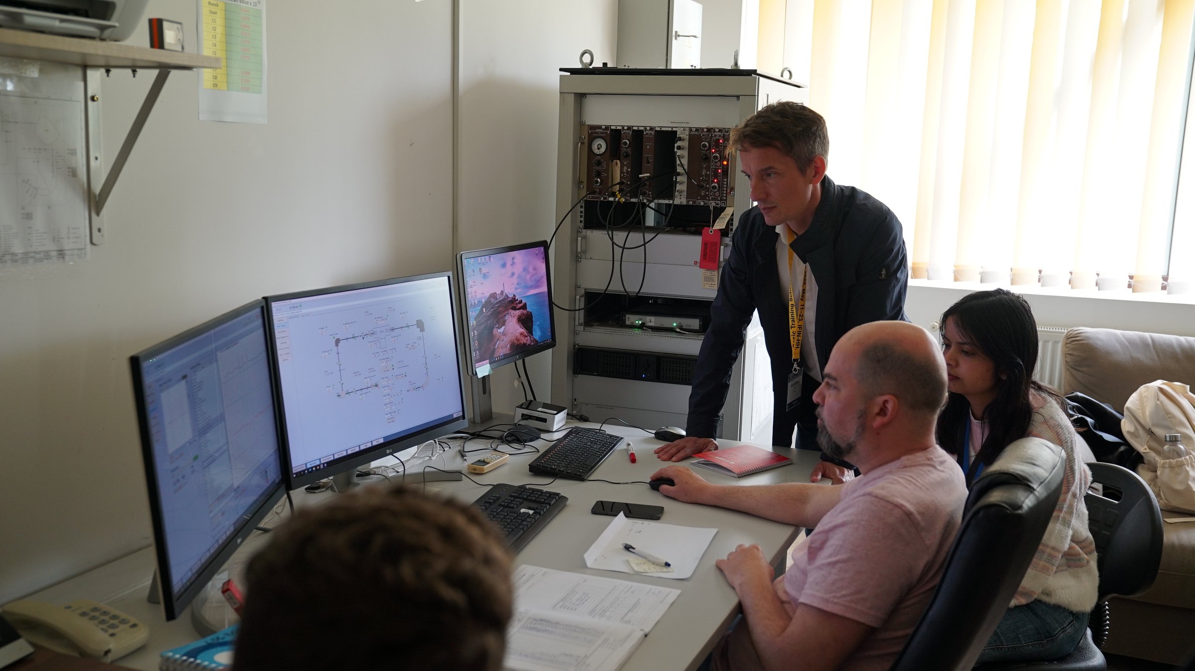 RoAMS operators at the control desk reading the beam-line control software on dual monitors, with the NIM electronics rack behind
