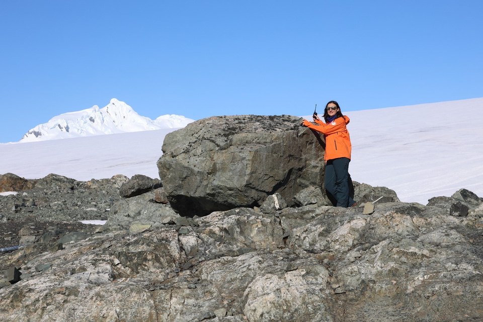Daniela Pascal beside a large boulder resting on bedrock at the edge of a snowfield