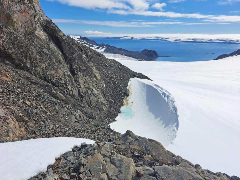 A rocky ridge beside a snowfield and glacier, with the sea and distant ice in the background