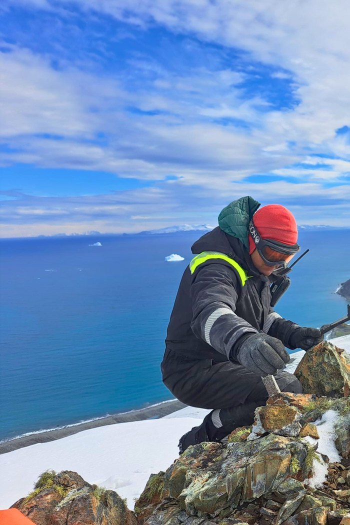 A researcher in cold-weather gear working on a rock outcrop on a ridge high above the sea