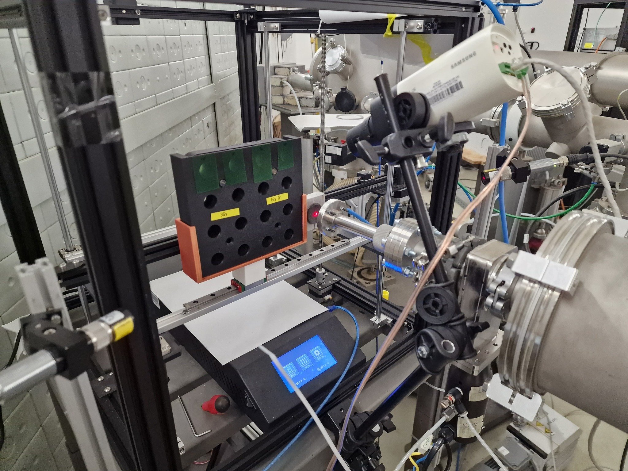 In-air irradiation station on the 3 MV proton beam line: a 3D-printed sample holder with dose-labelled wells (1 Gy, 3 Gy) positioned at the beam exit, with an XYZ stage and alignment camera